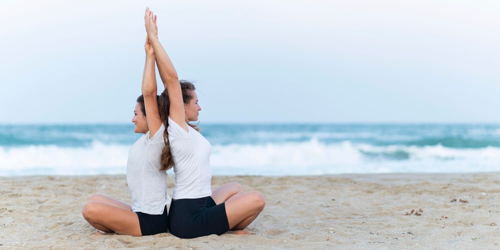 Two women practicing yoga back-to-back on a sandy beach with ocean waves in the background.
